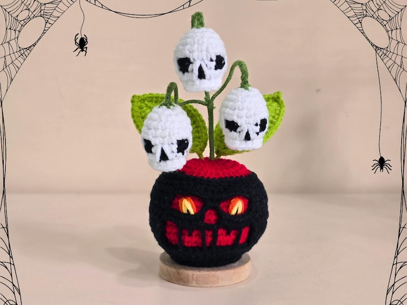 Peut inclure: D&eacute;coration d'Halloween en crochet repr&eacute;sentant une base noire et rouge en forme de citrouille avec des yeux orange brillants et une bouche souriante. Trois fleurs blanches en forme de cr&acirc;ne avec des feuilles vertes sortent du dessus.