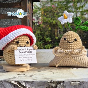 Peut inclure: Deux figurines de pommes de terre en crochet, l'une portant un chapeau de Père Noël rouge et tenant une pancarte sur laquelle est écrit "Emotional Support Santa Potato. I'm may be a tiny Santa Potato, but I believe in you. Go do your thing." L'autre figurine de pomme de terre a une tige verte et une fleur blanche.