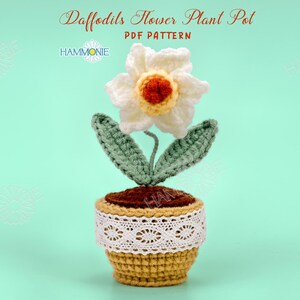 May include: Crochet pattern for a daffodil flower plant pot. The pot is brown with white lace trim and the flower is white with a yellow center and green leaves.