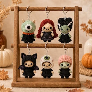 May include: Six crocheted keychains hanging on a wooden stand. The figures include a green one-eyed monster, a girl with red braided hair, a green-skinned character with black hair, a black bat, a white eyeball, and a pink-haired figure.