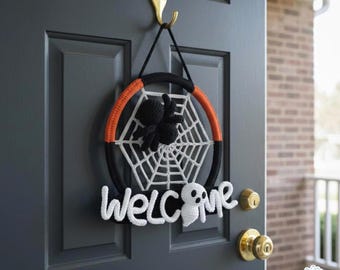 May include: A Halloween-themed door wreath with a black, orange, and white color scheme. The wreath features a spider web design with a black spider, and the word "WELCOME" in white. The wreath is hanging on a gray door.