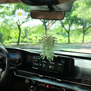 May include: A green crocheted plant hanging from the rearview mirror of a car. The plant is in a small white pot and has a long, trailing stem. The car's dashboard is visible in the background.