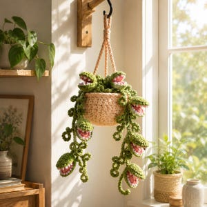 May include: A crocheted hanging plant featuring a light peach-colored pot and long, trailing green vines. The vines are adorned with multiple small, open-mouthed green and pink carnivorous plant figures with visible white teeth, suspended by a cord.