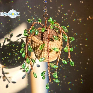 May include: A hanging, crocheted plant with green beads and copper wire. The plant is in a brown pot and has a rustic, handmade look.