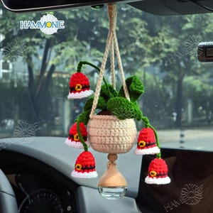 May include: A crocheted hanging planter with red and white bell-shaped flowers. The planter is hanging from a rearview mirror in a car. The planter is filled with green crocheted leaves and a small white crocheted pot.