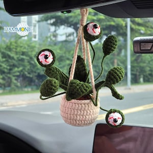 May include: A handmade crochet hanging plant with three eye-shaped flowers. The plant is in a beige pot and suspended by a beige rope. The eyes are white with red and black details. The brand name "HAMMONIE" is visible.