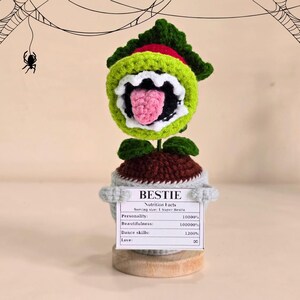 Peut inclure: Une plante crochet&eacute;e avec une grande bouche ouverte, des feuilles vertes et rouges, et une langue rose. La plante est dans un pot gris avec une pancarte "BESTIE" et des "Nutrition Facts". Toiles d'araign&eacute;es et araign&eacute;es en arri&egrave;re-plan.