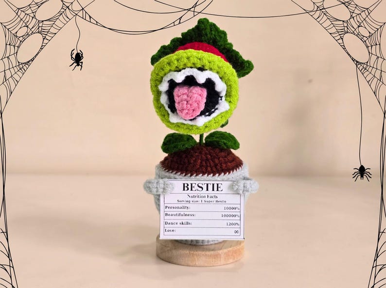 Peut inclure: Une plante crochet&eacute;e avec une grande bouche ouverte, des feuilles vertes et rouges, et une langue rose. La plante est dans un pot gris avec une pancarte "BESTIE" et des "Nutrition Facts". Toiles d'araign&eacute;es et araign&eacute;es en arri&egrave;re-plan.