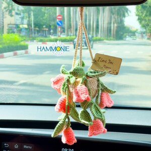 Op de afbeelding: Een gehaakt bloemstuk dat aan de achteruitkijkspiegel van een auto hangt. De bloemen zijn roze en wit, en de pot is beige. Een houten label met de tekst "Ik hou van je Mama" is aan de pot bevestigd.