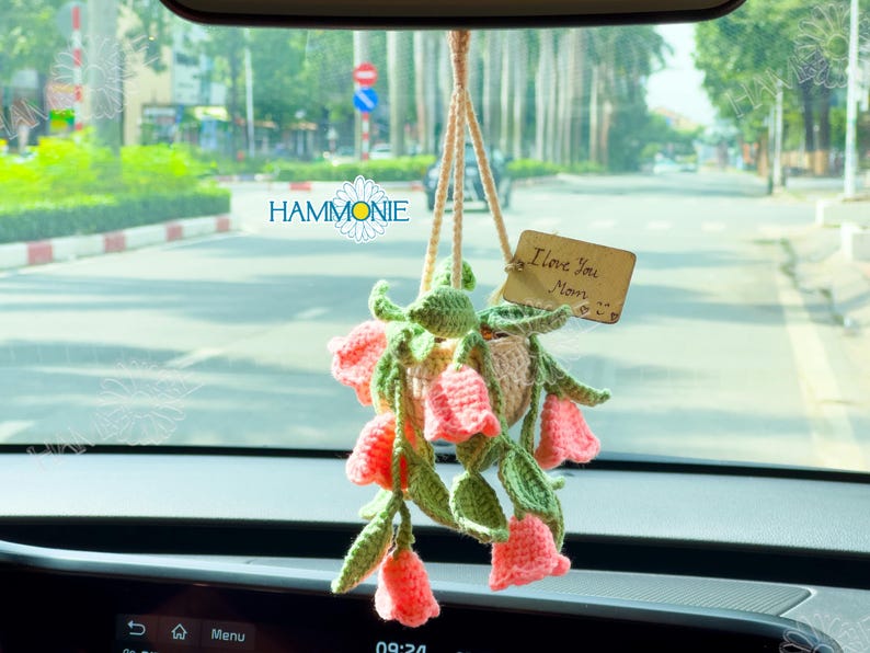 Op de afbeelding: Een gehaakt bloemstuk dat aan de achteruitkijkspiegel van een auto hangt. De bloemen zijn roze en wit, en de pot is beige. Een houten label met de tekst "Ik hou van je Mama" is aan de pot bevestigd.