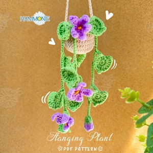 Könnte beinhalten: Eine gehäkelte Hängepflanze mit lila Blüten und grünen Blättern. Die Pflanze befindet sich in einem beigefarbenen Topf und hängt an einer Schnur. Der Text "Hanging Plant PDF Pattern" befindet sich am unteren Rand des Bildes.