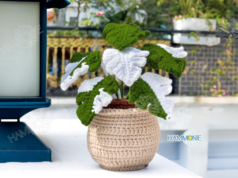 May include: A crocheted plant with white and green leaves in a beige crocheted pot. The plant is sitting on a white surface.