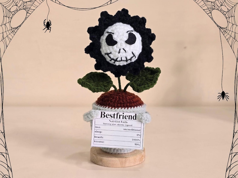 Peut inclure: Fleur au crochet avec un visage de cr&acirc;ne, des p&eacute;tales noirs et des feuilles vertes. La fleur est dans un pot avec une &eacute;tiquette indiquant "Bestfriend" et "Nutrition Facts". Le fond pr&eacute;sente des toiles d'araign&eacute;es et des araign&eacute;es.