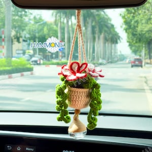 May include: A crocheted hanging planter with red and white flowers and green leaves. The planter is hanging from a rearview mirror in a car.