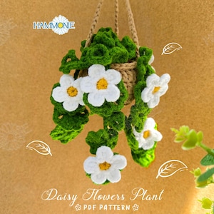 May include: A crocheted daisy flower plant hanging from a string. The plant is in a brown pot and has white flowers and green leaves. The background is a light brown.