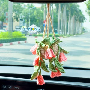 Op de afbeelding: Een gehaakte hangende bloempot met roze bloemen en groene bladeren. De pot hangt aan een achteruitkijkspiegel in een auto.