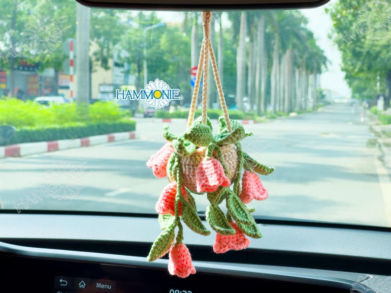 Op de afbeelding: Een gehaakte hangende bloempot met roze bloemen en groene bladeren. De pot hangt aan een achteruitkijkspiegel in een auto.