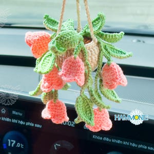Op de afbeelding: Een gehaakte hangende plantenpot met roze bloemen en groene bladeren. De pot hangt aan een achteruitkijkspiegel in een auto.
