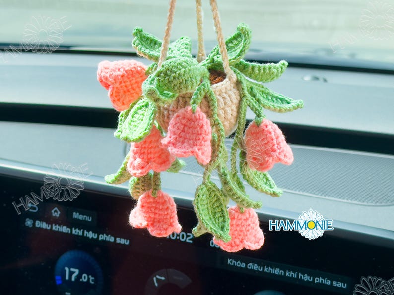 Op de afbeelding: Een gehaakte hangende plantenpot met roze bloemen en groene bladeren. De pot hangt aan een achteruitkijkspiegel in een auto.