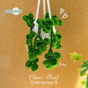 May include: A crocheted clover plant hanging from a white string. The plant is green and has a white pot. The background is a brown surface with a white daisy.