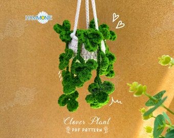 May include: A crocheted clover plant hanging from a white string. The plant is green and has a white pot. The background is a brown surface with a white daisy.