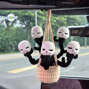 May include: A beige crochet car charm featuring five white skull heads with black accents. The skulls are attached to black pipe cleaner arms and are suspended from a beige woven basket. The charm hangs from a beige cord.