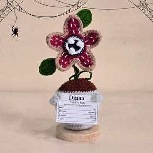 Peut inclure: Fleur de Demogorgon crochet&eacute;e avec une fleur bordeaux et beige, des feuilles vertes et un centre blanc et noir. La fleur est pos&eacute;e sur un pot marron et gris avec une &eacute;tiquette indiquant "Diana Nutrition Facts". La base est un disque en bois.