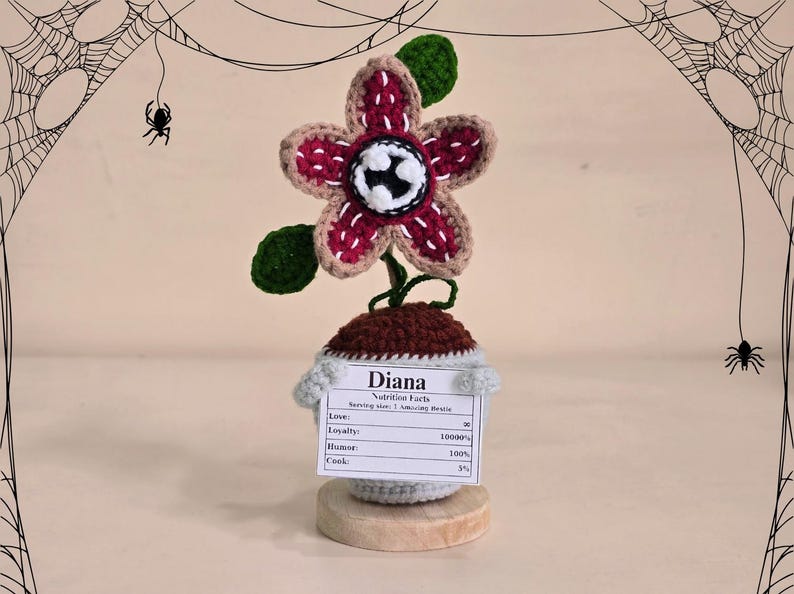Peut inclure: Fleur de Demogorgon crochet&eacute;e avec une fleur bordeaux et beige, des feuilles vertes et un centre blanc et noir. La fleur est pos&eacute;e sur un pot marron et gris avec une &eacute;tiquette indiquant "Diana Nutrition Facts". La base est un disque en bois.