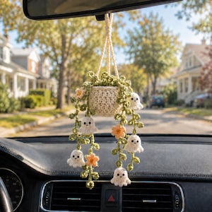 May include: A crocheted hanging plant ornament featuring a cream-colored pot, green vines, small orange flowers, and several tiny white ghost figures with black eyes and mouths, suspended by a cream-colored cord from a vehicle rearview mirror.
