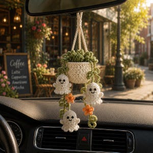 May include: A crocheted car accessory featuring a small cream-colored pot holding green vines with three white ghost figures and two orange flowers. The item is suspended by a cream-colored cord, designed to hang from a vehicle rearview mirror.