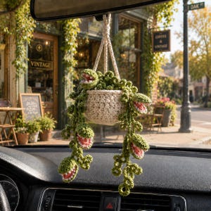 May include: A crocheted hanging plant ornament featuring a cream-colored pot and trailing green vines with several open, toothy, pink-centered carnivorous plant heads. The item is suspended by a braided cord, designed for display in a vehicle.