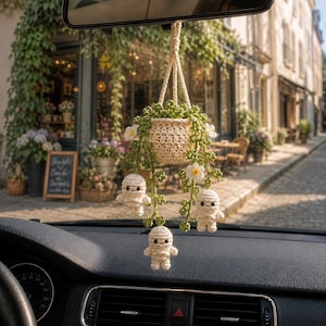 May include: A crocheted car hanging ornament featuring a cream-colored pot holding a green trailing plant with small white flowers and three small, cream-colored, wrapped mummy figures dangling from the vines by thin crocheted cords.