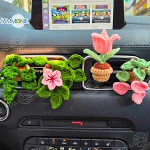 May include: Four crocheted car air freshener decorations. Two are green and pink flower arrangements, one is a pink tulip, and one is a green plant. All are in brown pots.
