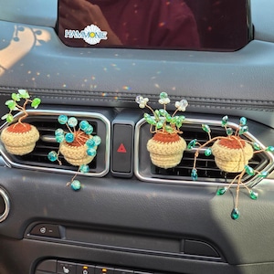 May include: Four crocheted potted plants with green and blue beads hanging from the car's air vents. The plants are in brown pots with green leaves and white flowers.