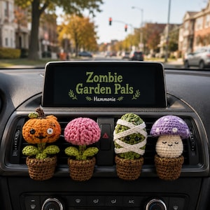 May include: Four crocheted plant ornaments attached to a car air vent. The figures include an orange pumpkin with button eyes, a pink flower, a green cactus wrapped in white bandages, and a purple mushroom with white spots, all in brown pots.