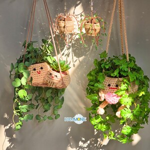 May include: Two hanging plant pots with smiling faces. The pots are made of crocheted yarn and are filled with green plants. The pots are hanging from the ceiling by brown cords.