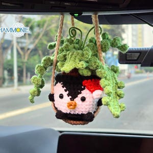May include: A crocheted penguin wearing a red Santa hat is holding a green plant in a pot. The penguin and plant are hanging from the rearview mirror of a car.