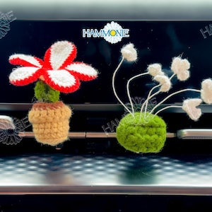 May include: Two crocheted car air freshener ornaments. One is a red and white flower in a brown pot. The other is a green pot with white flowers.