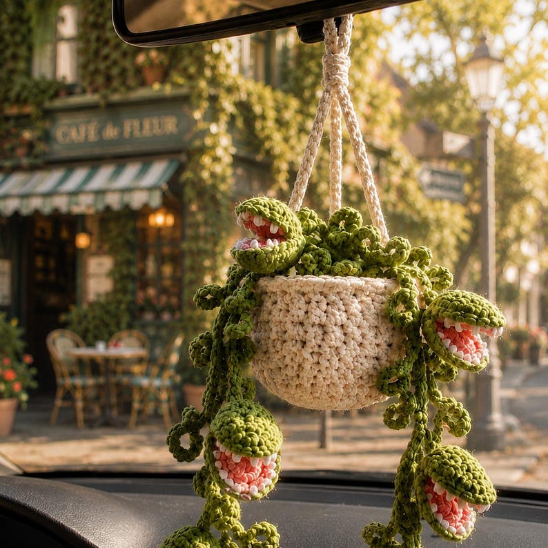 May include: A crocheted hanging plant ornament featuring a cream-colored pot and several green, vine-like stems with open, toothy, pink-centered mouths. The decorative piece is suspended by a braided cord, designed for display in a vehicle interior.