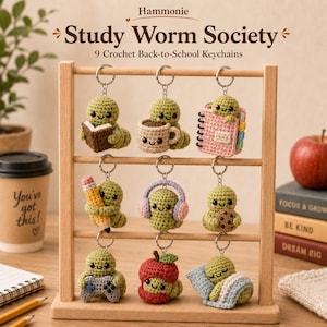May include: Nine crochet keychains featuring green worms with smiling faces, displayed on a wooden rack. The worms are depicted with various accessories including a book, coffee mug, notebook, pencil, headphones, cookie, game controller, apple, and blanket.