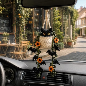 May include: A crochet car accessory featuring a black cat head peeking out of a cream-coloured pot. The cat has bright yellow eyes. Trailing down from the pot are green vines with small black bat shapes and vibrant orange flowers attached.