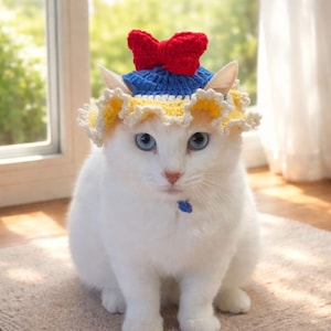 Op de afbeelding: Een witte kat draagt een gehaakte hoed met een blauwe kroon, een rode strik bovenop en een brede, geplooide gele rand met een witte geschulpte afwerking. De hoed is zo ontworpen dat deze rond de oren van de kat past.