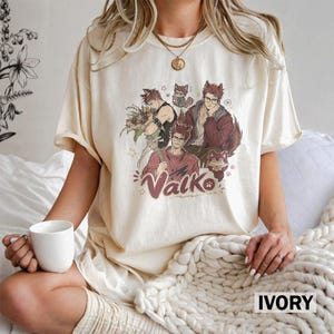 May include: An ivory-colored short-sleeve t-shirt featuring a graphic print of three anime-style characters with wolf ears and tails. The text "Valko" is printed in a stylized red font below the characters, accompanied by small star and paw print icons.