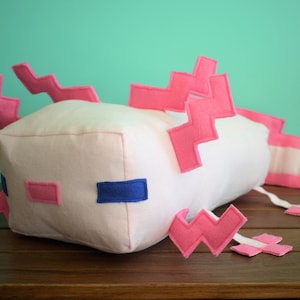 May include: A white plush axolotl toy with pink felt accents. The toy has a square body and a long tail with pink felt accents. The toy is lying on a wooden surface.