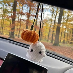 May include: A crochet car hanging ornament featuring a small orange pumpkin with a green stem and a white ghost with black bead eyes and pink cheeks, both suspended from a black string against a blurred background of autumn trees.