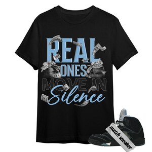 May include: A black short-sleeve t-shirt featuring the light blue text REAL ONES and the white text MOVE IN, with the word Silence in light blue script. The design includes illustrations of black money bags and floating dollar bills.