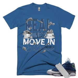 May include: A royal blue short-sleeve t-shirt featuring the text "REAL ONES MOVE IN SILENCE" in white and blue lettering, accompanied by graphic illustrations of two white money bags overflowing with cash and floating dollar bills.