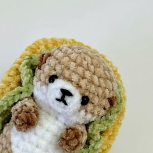 Op de afbeelding: Een kleine, gehaakte pluche otter met een beige en wit lichaam, zwarte kraaloogjes en een geborduurde zwarte neus. De otter ligt in een geel en groen gehaakte structuur die lijkt op een taco of een opgevouwen deken.
