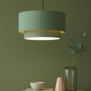 Op de afbeelding: Een tweeledige, saliegroene hanglamp hangt aan het plafond. De lamp hangt boven een houten bijzettafel met een vaas, een kom en een roze vaas. De achtergrond is effen olijfgroen.