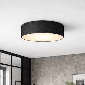 Könnte beinhalten: Eine runde Deckenleuchte aus schwarzem Stoff. Die Leuchte hat einen zylindrischen Schirm und einen weißen Innenraum, der für eine weiche, umgebende Beleuchtung sorgt. Die Leuchte ist an einer weißen Decke montiert, passend für verschiedene Einrichtungsstile.